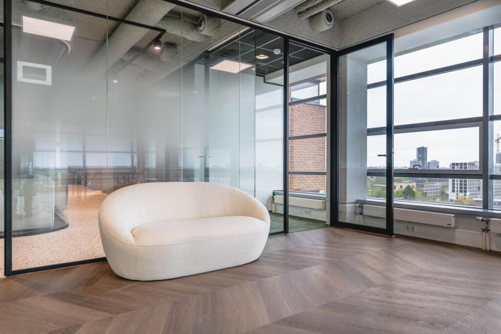 Modern office space at Joop Geesinkweg with a white curved sofa, glass walls, and city view through large windows.